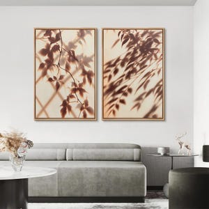 May include: Two rectangular wall art pieces in light wood frames, each displaying a sepia-toned photograph of foliage shadows cast against a grid pattern. The left print shows distinct leaf silhouettes, while the right print features elongated, drooping branch shadows.