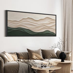 May include: Abstract wall art featuring a layered landscape with beige and green hues. The artwork is framed in black and hangs on a white wall above a brown couch.