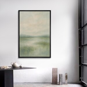 May include: A framed landscape painting with a black border. The artwork features a serene scene with muted green and beige tones, depicting a body of water and distant hills under a soft sky. The painting evokes a sense of calm and tranquility.