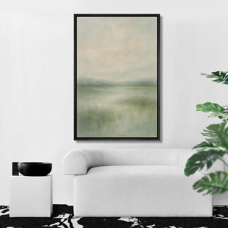 May include: A framed landscape painting with a serene, muted color palette of greens, beige, and gray. The artwork depicts a misty lake or marsh scene, with soft brushstrokes creating a sense of depth and tranquility. The black frame complements the artwork's minimalist aesthetic.