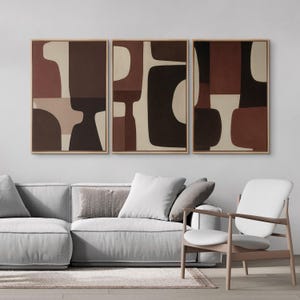 May include: Triptych of abstract art prints. Each framed artwork features a design of geometric shapes in shades of brown, beige, and black. The artwork is displayed above a gray sofa and a white chair.
