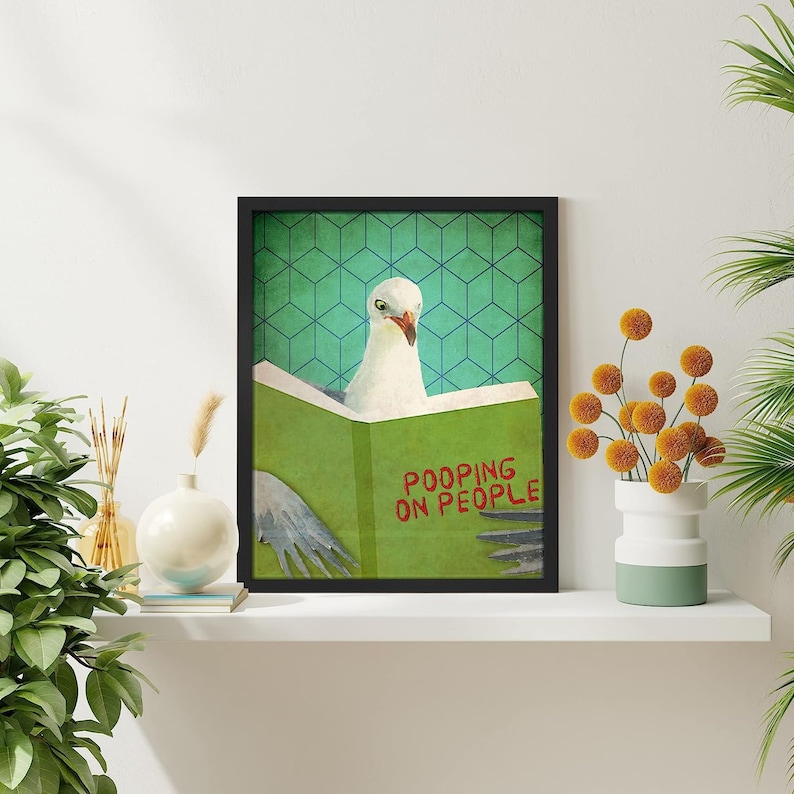 K&ouml;nnte beinhalten: Ein gerahmter Druck einer M&ouml;we, die ein Buch mit dem Titel "Pooping on People" liest. Das Buch ist gr&uuml;n und die M&ouml;we ist wei&szlig;. Der Hintergrund ist ein t&uuml;rkisfarbenes und gr&uuml;nes geometrisches Muster.
