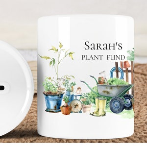 Könnte beinhalten: Weiße Keramik-Sparbüchse mit einem Schlitz oben. Die Vorderseite der Sparbüchse zeigt eine Aquarell-Illustration von Gartengeräten, Pflanzen und einer Schubkarre. Der Text "Sarah's Plant Fund" ist in Schwarz auf der Vorderseite der Sparbüchse aufgedruckt.