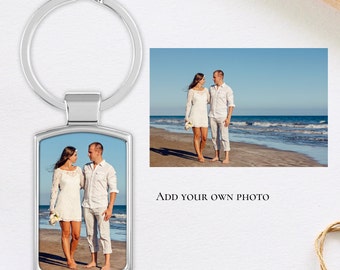 Puede incluir: Un llavero plateado con un inserto de foto rectangular que muestra a una pareja con ropa blanca caminando por una playa. El texto "ADD YOUR OWN PHOTO" está debajo de la imagen.