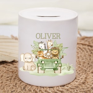 May include: White ceramic money box with a slot on top, decorated with a safari scene. The design includes a jeep filled with animals like a lion, tiger, zebra, and giraffe, with the name "OLIVER" above. The bank is cylindrical in shape.