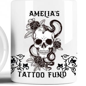 Pode incluir: Caneca de cerâmica branca com um desenho de caveira a preto e branco, uma cobra e rosas. O texto "AMELIA'S TATTOO FUND" é impresso na caneca com uma fonte gótica. A caneca tem uma borda preta decorativa.