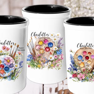 May include: Three white ceramic mugs with black rims, each featuring a watercolor design of a paint palette with colorful paint, paintbrushes, and flowers. The text "Charlotte's Paint Brushes" is printed on each mug.