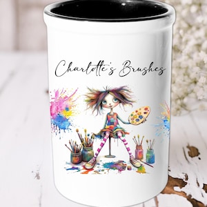 Puede incluir: Portacepillos de cerámica blanca con borde negro. El portacepillos presenta una ilustración colorida de una niña con salpicaduras de pintura y el texto "Charlotte's Brushes".