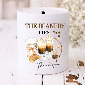 Könnte beinhalten: Eine weiße Keramik-Tippdose mit dem Text "The Beanery Tips" und einer Grafik von zwei Kaffeegetränken mit Schlagsahne, einem Croissant und einem Cupcake. Der Text "Thank you" steht unter der Grafik.