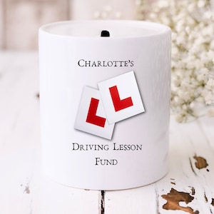 Op de afbeelding: Witte keramische spaarpot met een gleuf aan de bovenkant. Hij heeft twee rode "L" erop gedrukt en de tekst "CHARLOTTE'S DRIVING LESSON FUND".