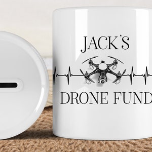 Peut inclure: Tirelire en céramique blanche avec une fente sur le dessus. Le devant de la tirelire présente une image en noir et blanc d'un drone avec une ligne de battement de cœur et le texte "JACK'S DRONE FUND".