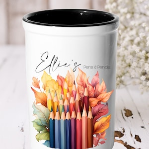 May include: White ceramic pencil holder with a black rim. It features a colourful illustration of pencils and autumn leaves with the text "Ellie's Pens & Pencils".