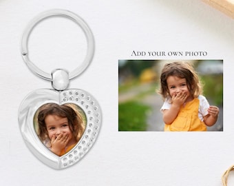 May include: Silver heart-shaped keychain with a photo frame. The frame contains a photo of a young girl smiling and covering her mouth with her hand. The keychain has a silver ring for attaching to keys.