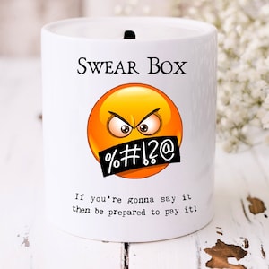 Könnte beinhalten: Eine weiße Keramik-Schimpfwort-Dose mit schwarzem Deckel. Die Dose hat ein gelbes Emoji-Gesicht mit einem schwarzen Zensurbalken über dem Mund. Der Text auf der Dose lautet "SWEAR BOX" und "If you're gonna say it then be prepared to pay it!"