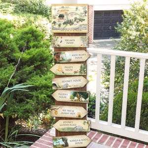 May include: A wooden signpost with multiple arrows pointing to different locations in the Hundred Acre Wood. Each arrow features a character from Winnie the Pooh, including Pooh Bear, Piglet, Eeyore, Tigger, and Kanga and Roo. The signpost is decorated with illustrations of the characters and their homes.