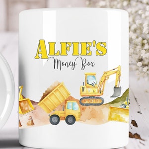 May include: White ceramic money box featuring a yellow and black construction vehicle design. The text "Alfie's Money Box" is printed on the front of the mug.