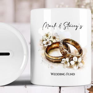 May include: White ceramic wedding fund bank with a slot on top. The bank features a watercolor design of two gold wedding rings, white flowers, and the text "Mark & Stacey's" and "Wedding Fund."