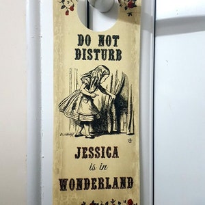 Op de afbeelding: Een vintage-stijl deurhanger met een zwart-witte illustratie van Alice uit Alice in Wonderland die achter een gordijn vandaan kijkt. De tekst luidt "Niet storen Jessica is in Wonderland".