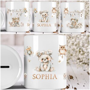 Peut inclure: Tirelire en céramique blanche sur le thème de la forêt. La tirelire représente un ours brun portant un chapeau, un oiseau bleu, un hibou et le nom "SOPHIA" en lettres dorées.
