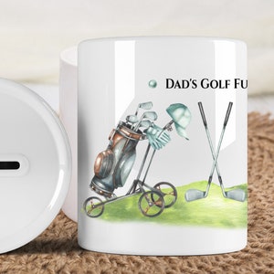 May include: White ceramic mug with a golf-themed design. The illustration features a golf bag on a cart, golf clubs, a golf hat, and the text "Dad's Golf Fun". A white piggy bank is in the background.