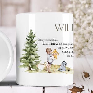 Könnte beinhalten: Weiße Keramiktasse mit einer Winnie-the-Pooh-Illustration. Die Tasse zeigt einen Jungen, der einen Teddybär hält, einen Baum und ein Zitat, das lautet: "Denke immer daran... Du bist MU-TIGER als du glaubst, STÄRKER als du scheinst und KLU-GER als du denkst." Der Name "WILLI" ist oben auf der Tasse aufgedruckt.