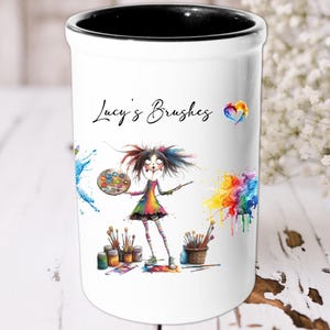 Puede incluir: Portaútiles de cerámica blanca con borde negro. Presenta una ilustración colorida de una niña con salpicaduras de pintura y el texto "Lucy's Brushes" con un corazón arcoíris.