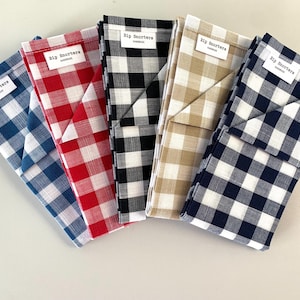 Op de afbeelding: Vijf gevouwen stoffen items in blauw, rood, zwart, beige en marine, elk met een ruitpatroon. Ze hebben een klein wit label met de tekst "Rip Snorters handmade".