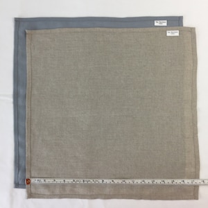 May include: Two linen napkins, one light gray and one beige, with a measuring tape placed along the bottom edge. The beige napkin has a small white tag with the text "Rip Stop Works" on it.