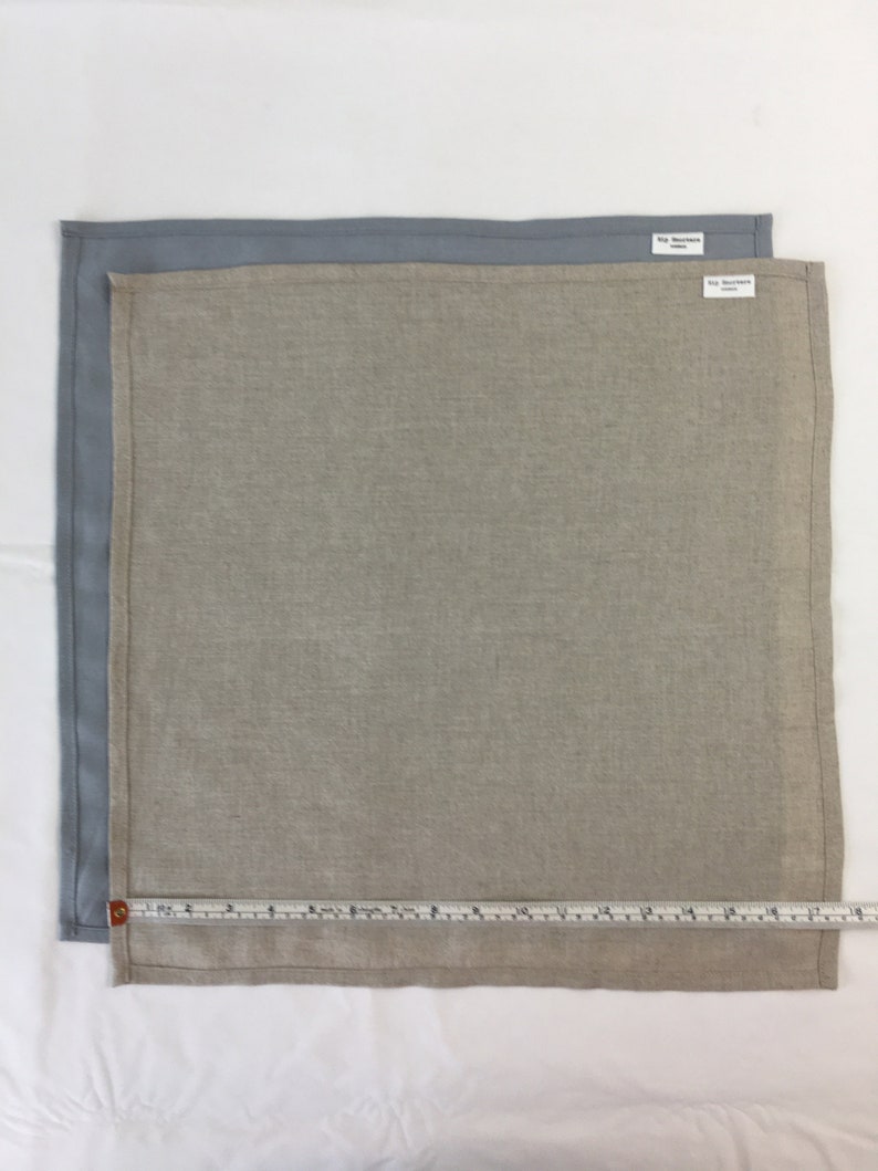 May include: Two linen napkins, one light gray and one beige, with a measuring tape placed along the bottom edge. The beige napkin has a small white tag with the text "Rip Stop Works" on it.