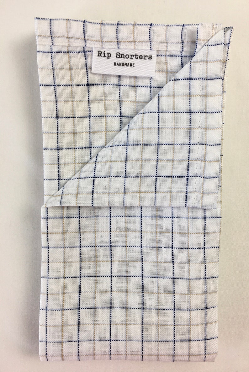 May include: A white kitchen towel with a blue and tan plaid pattern. The towel is folded in half and has a label that reads "Rip Snorters Handmade".