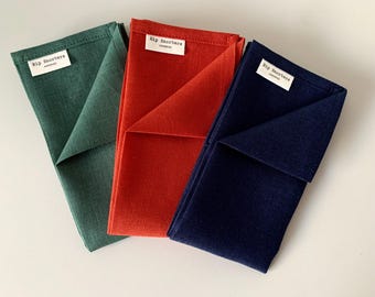May include: Three folded fabric napkins in forest green, burnt orange, and navy blue. Each napkin features a white rectangular fabric label sewn near the top edge with the black text Rip Snorters HANDMADE printed on the surface of the label.