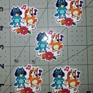 May include: A collection of stickers featuring cartoon characters. Each sticker shows two cartoon dogs, one blue and one orange, standing on a cloud with flowers. The blue dog wears headphones, and both have musical notes.