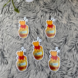May include: Five stickers featuring a cartoon bear in a red shirt sitting in a honey pot labeled "HUUNY". The stickers are surrounded by small, stylized bees. The background is a gray, textured surface.