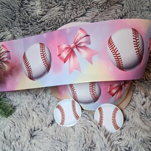 May include: A baseball-themed ribbon with a pastel background featuring baseballs and pink bows. Two baseball-shaped buttons are also visible. The ribbon and buttons are likely for crafting or decorative purposes.