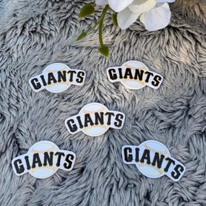 May include: Five baseball-themed stickers with the word "GIANTS" in black lettering. Each sticker features a baseball design with red stitching. The stickers are on a gray, textured surface, with white flowers in the background.