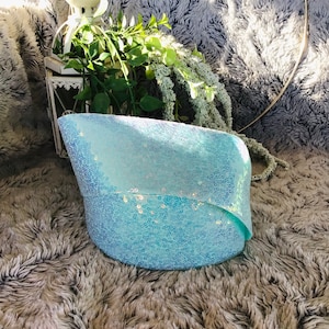 Puede incluir: Una almohada azul claro con lentejuelas y un ligero brillo iridiscente. La almohada está doblada por la mitad y se encuentra sobre una superficie gris y esponjosa.