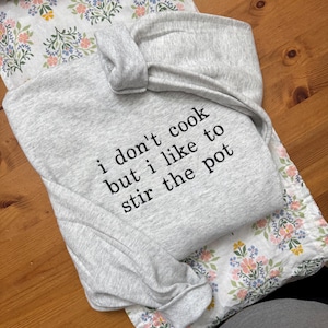 May include: A light gray sweatshirt with the text "i don't cook but i like to stir the pot" in black. The sweatshirt is folded on top of a floral patterned pillowcase. The background is a wooden surface.