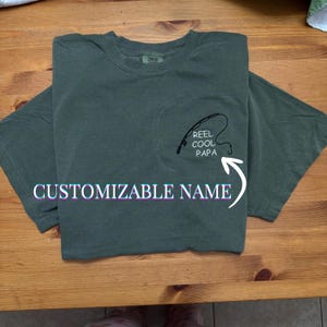 Może przedstawiać: Zielona koszulka z kieszonką z czarnym wzorem wędki i napisem "REEL COOL PAPA". Koszulka ma również napis "CUSTOMIZABLE NAME" pod kieszenią.