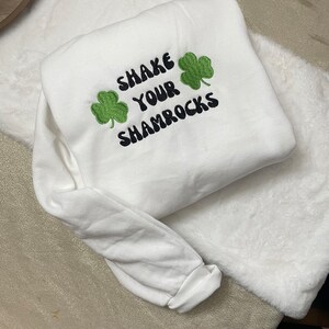 May include: White sweatshirt with the text "Shake Your Shamrocks" embroidered in black with two green shamrock designs.