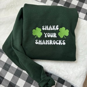May include: Green sweatshirt with white embroidered text that says "Shake Your Shamrocks" with two green shamrock designs on either side of the text.
