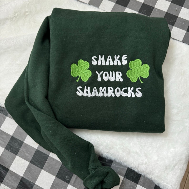 May include: Green sweatshirt with white embroidered text that says "Shake Your Shamrocks" with two green shamrock designs on either side of the text.