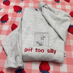 May include: A light grey sweatshirt featuring an embroidered duck behind bars, with the red text "got too silly" beneath. The sweatshirt is folded on a pink and white strawberry-patterned surface.