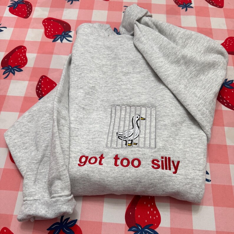 May include: A light grey sweatshirt featuring an embroidered duck behind bars, with the red text "got too silly" beneath. The sweatshirt is folded on a pink and white strawberry-patterned surface.