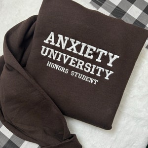 May include: A brown sweatshirt with the text "Anxiety University Honors Student" embroidered in white.