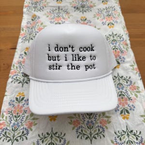 Op de afbeelding: Witte truckercap met zwarte geborduurde tekst die "ik kook niet, maar ik vind het leuk om de pot te roeren" zegt.