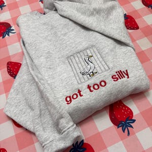 May include: A grey hoodie with a hood and long sleeves. The front has an embroidered design of a duck behind bars and the phrase "got too silly" in red thread. The hoodie is folded on a pink and white checkered surface with strawberry designs.