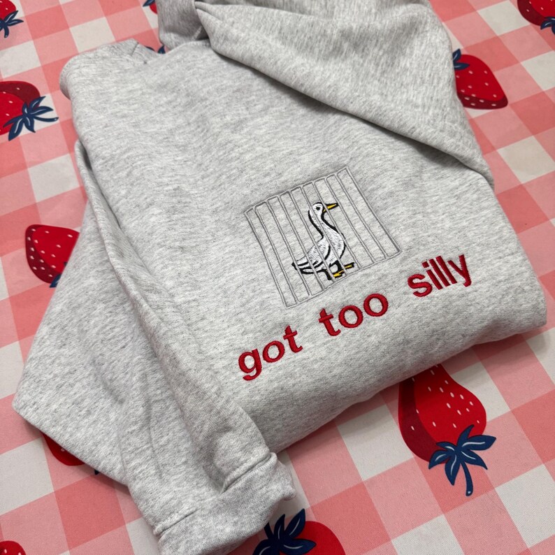 May include: A grey hoodie with a hood and long sleeves. The front has an embroidered design of a duck behind bars and the phrase "got too silly" in red thread. The hoodie is folded on a pink and white checkered surface with strawberry designs.