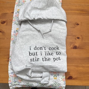 Könnte beinhalten: Grauer Kapuzenpullover mit dem Aufdruck "i don't cook but i like to stir the pot" auf der Vorderseite.