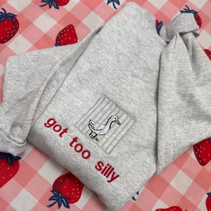May include: A light grey sweatshirt with the text "got too silly" in red. It has an embroidered patch of a goose in a cage. The background is a pink and white gingham pattern with strawberry motifs.