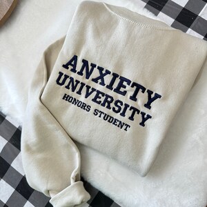 May include: A beige sweatshirt with the text "Anxiety University Honors Student" embroidered in navy blue.